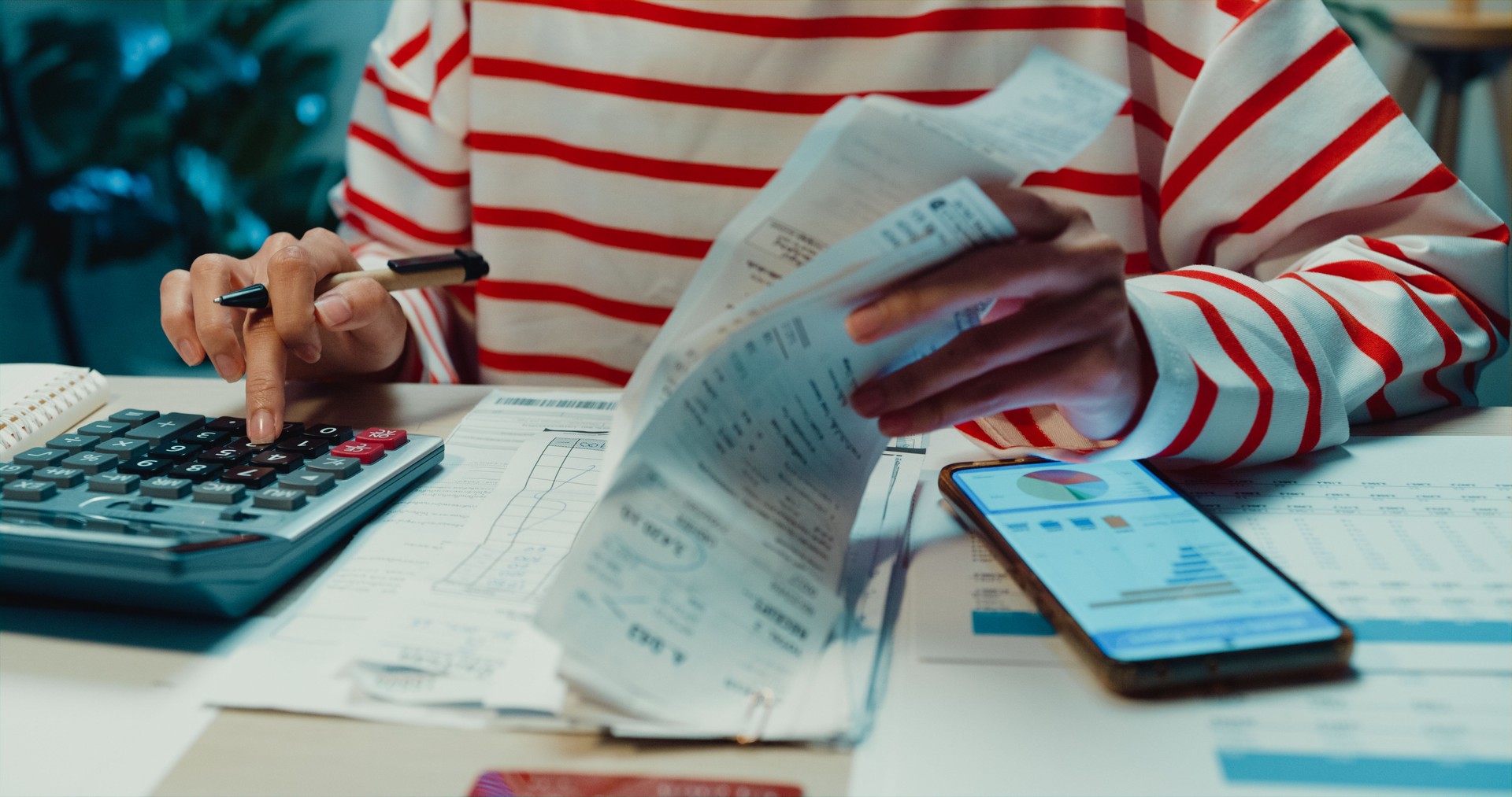 Close-up Young Asia woman with sweater sit in front of desk with phone use calculator to calculate utility bills check credit card receipt monthly expense bill in house at night.